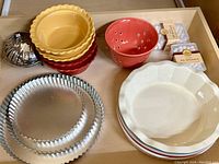 Overview of ceramic bakeware and some kitchen tools in drawer, including scalloped edge pie dishes in yellow, red, and cream colors, metal steamer basket, ceramic colander, and pie weight boxes