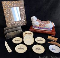 Full assortment of vintage Asian decorative items including gilt gesso mirror, cloisonné letter holder, porcelain head rest, small dish, coasters, combs, and carved hairpiece arranged on black backdrop.