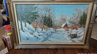 Full view of the framed oil painting showing a winter scene with a cottage and snow-covered ground under a blue sky.