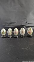 Five vintage decorative eggs with animal and nature designs, each on black wooden stands, arranged in a row against a black backdrop.