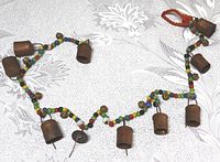 Photo showing the entire string with seven visible large coppery metal bells, colorful glass beads, and small brass bells on a light floral-patterned tablecloth.