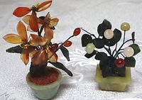 Two carved stone miniature tree figurines displayed on a white patterned cloth; one orange stone flower in green pot, one with stone fruits on square base missing one fruit