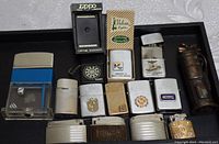 Full view of 14 assorted vintage cigarette lighters arranged on a tray, showing various brands and designs including dice lighter, dartboard Zippo, Vulcan box lighter.
