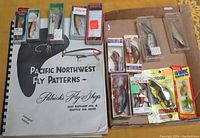 Lot laid out on surface showing fishing lures mostly in packaging and a fishing patterns book.