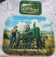 Two John Deere tin trays, one large with a farm scene and green tractor, one smaller with tractor in front of barn