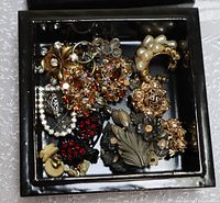 Top view of black lacquer square box filled with assorted costume jewelry including earrings and brooches.