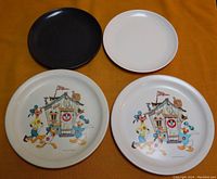 Four plastic plates on an orange background. Two 9-inch Mickey Mouse Club decorative plates with cartoon characters and two smaller plain black and white plastic plates.