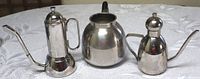 Front view of three stainless steel coffee and tea pots showing different styles and sizes on a white patterned tablecloth.