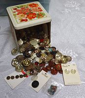 Open flower-themed tin container showing a large quantity of assorted vintage buttons spilling out, along with two cloisonné owl thimbles and button cards