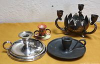 Four candleholders shown together on yellow background including shiny silverplate chamberstick, old pewter chamberstick, small decorative Noritake ceramic candleholder, and tarnished silverplate centerpiece with flower bowl.