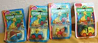 Four Sesame Street die cast vehicles still sealed in original blister packaging. Vehicles include a blue and brown Snuffle-upagus dinghy, blue and white Big Bird mail truck, yellow and red Cookie Monster express train, and green and yellow Oscar the Grouch trash delivery truck. Packaging features colorful Sesame Street branding with character images.