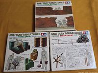 Three boxed Tamiya 1/35 scale military miniature model kits placed on wooden surface. Boxes show illustrations of items included: barricades, brick walls, and jerry cans with figures.