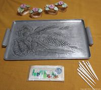 Aluminum tray with embossed fruit and grain design, ceramic floral napkin holders, carved appetizer picks, and packaged German plastic napkin clips shown arranged on yellow fabric.