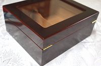 Close-up photo showing the reddish-brown wooden humidor box with glass lid and brass hinges.