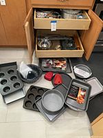 Wide view showing assortment of metal muffin pans, rectangular and square baking pans, bundt cake pan, glass bowls, red silicone oven mitts, all laid on floor in front of open cabinet drawers.