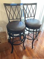 Front view of the pair of metal and leather barstools placed side by side on wooden floor, showing decorative backrest and round leather padded seats.