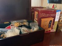 Open wooden jewelry chest holding costume jewelry in plastic bags, visible gold-tone brooch with filigree design.