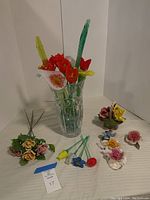 Full view of glass vase with glass flowers and painted porcelain flowers arranged around it on table.