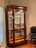 Front view of the wooden frame glass china cabinet with interior shelves and light, sitting next to a window and chair.