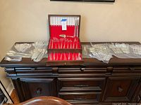 Full overview of the silverware set laid out on a wooden cabinet, showing the flatware pieces in their plastic sheathes and the wooden storage box.