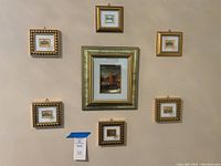 Photo of seven small framed wall art pieces arranged on a wall in gold leaf ornate frames, depicting classic European street and landmark scenes.
