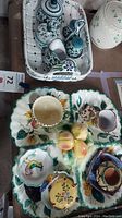Top view of ceramic basket tray holding salt and pepper shakers, creamer, sugar bowl, and toothpick holder with floral patterns. 4-section floral tray with painted fruits and roosters in front.