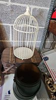 Photo showing cream-colored metal decorative birdcage on top of wooden picnic basket and green painted wooden firkin in front.