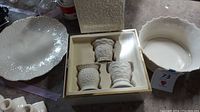 View of large white ornate serving plate and white scalloped edge bowl alongside boxed votive holders set