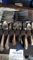 Photo showing an organized set of Oneida flatware with knives, forks, spoons grouped in rows. Pieces show surface tarnish and wear.