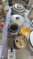 Overview of pottery lot items on table including covered baking dishes, floral jug, tart warmer, and crock.