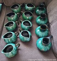 Photo showing 13 assorted blue-green glazes creamers by Blue Mountain Potteries with various shapes and spouts.