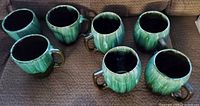 Seven green ceramic mugs arranged on a couch, showing color gradient glaze and rounded rims.
