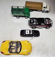 Five die cast vehicles arranged on a light surface showing two trucks, one police car, one black racing car, and one yellow convertible sports car.