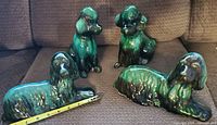 Four glazed ceramic dog figurines on a brown couch: two sitting poodles and two reclining Afghan hounds. Green and black coloration with glossy finish.