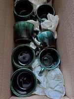 Top view of four coffee mugs inside a cardboard box, showing their dark green flowing glaze interior and exterior.