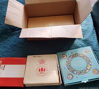 Four collectible plate boxes arranged on a blue quilt: two boxes with 12 Days of Christmas plates, one Villeroy & Boch Christmas in Europe plate box, and one Bing & Grondahl 1980 plate box.