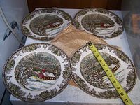 Four 10-inch dinner plates displayed with a tape measure showing size. Plates decorated with 'The Old School House' winter scene by Johnson Bros.