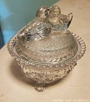 Pressed glass footed dish with two birds on lid as handle, standing on three feet with intricate patterns.