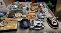Wide view of entire assortment laid out on a surface showing variety of collectible small items including belt buckles, metal car logos, spoons, medallions, and novelty items.