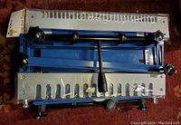 Top-down view of blue dovetail jig and two aluminum template plates wrapped in plastic on carpet.