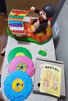 Photo showing monkey toy seated playing a colorful xylophone with 4 circular music disks and a small broken green lever lying next to it