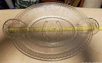 Oval glass serving platter with horseshoe handles shown with measuring tape to show length approximately 19 inches.
