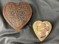 Two heart-shaped boxes placed side by side on a dark fabric background, one wooden with brass floral design and the other smaller stone with mother of pearl inlay.