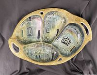 Top view of the oval ceramic serving dish showing four divided sections with abstract painted cityscape artwork inside and two cutout handles. The textured golden-brown exterior is visible with small cutouts near the handles.