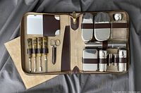 Grooming travel kit open inside leather and suede case showing all items in organized slots including manicure tools, mirror, containers, brushes, scissors, and aftershave bottle