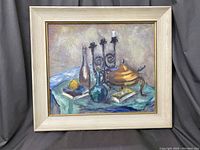 Framed oil painting depicting a still life with glass bottles, a lidded dish, books, candle holder, and lemon on a cloth surface.