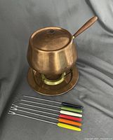 Copper pot with wood handle on brass stand with drip plate, and six fondue forks arranged in front