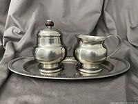 Front view of the sugar bowl with lid and creamer pitcher on the tray, showing the rounded stainless steel design and dark lid knob.