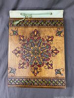 Front cover of vintage photo album showing wooden decorative cover with colorful floral and geometric hand-painted design, green braided cord with tassel.