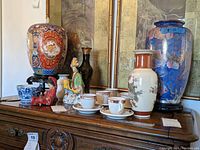 Photo of a collection of various Asian decorative items on a wooden surface, including ginger jars, vases, porcelain cups and saucers, ceramic figures, and a paper fan.